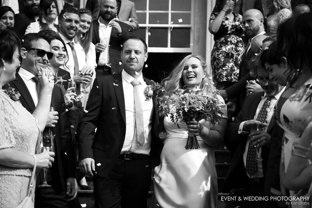 Black and white shot of a bride and groom making their way down the steps at their Kelmarsh Hall wedding as guests throw confetti