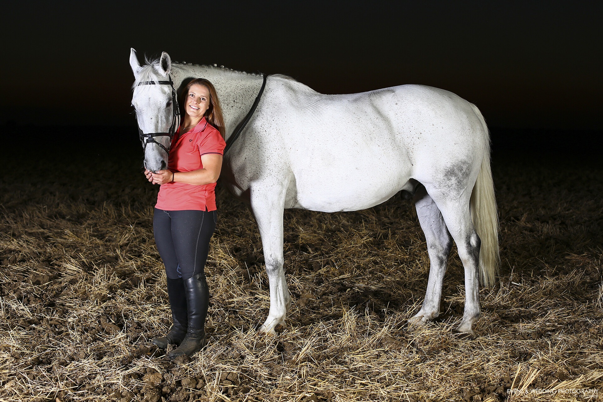 Horse and female rider stood in a ploughed field after dark, illuminated by studio lights