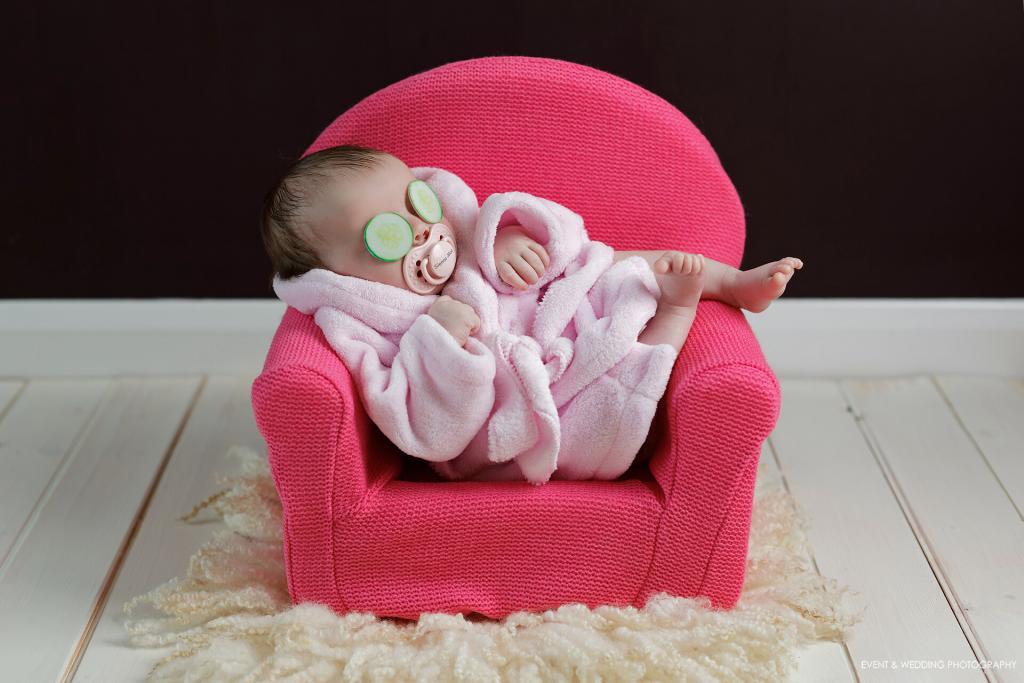 Baby girl asleep, wearing a dress gown, cucumber slices on her eyes, laying back in a shocking pink woollen newborn sofa