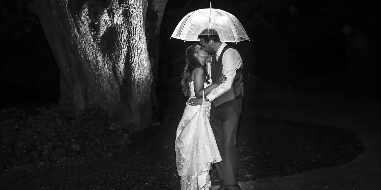 After dark black and white shot of the bride and groom underneath an umbrella in the rain at their Cranford Hall wedding.