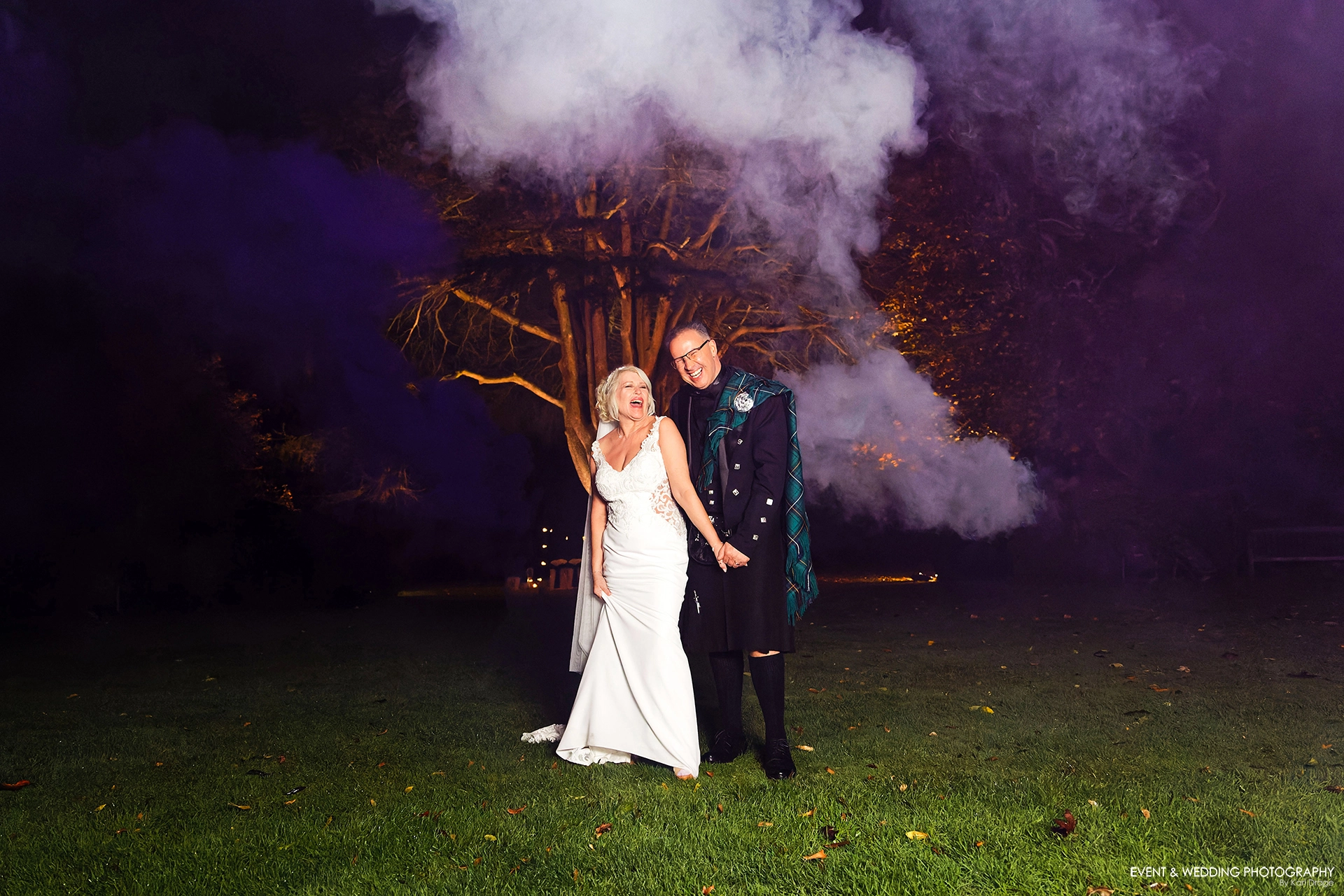 A bride and groom share a joke after dark in the grounds of Ansty Hall with purple and white smoke behind them