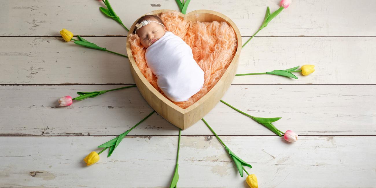 A newborn baby girl asleep in a heart-shaped wooden photo prop