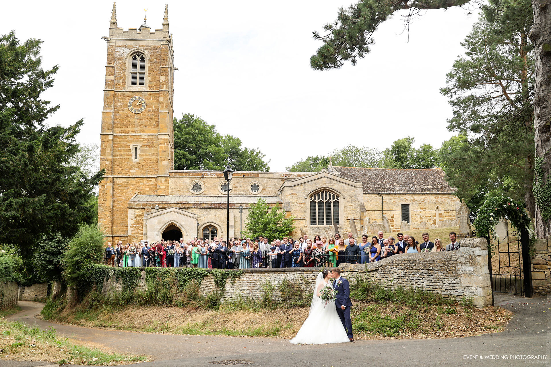 The bride and groom share a kiss on their Gretton Church wedding day as all of their guests line up against the railings, with the church providing the backdrop.