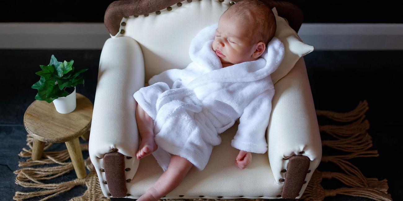 Newborn baby boy wearing a white dressing gown asleep on a white and brown upholstered sofa.