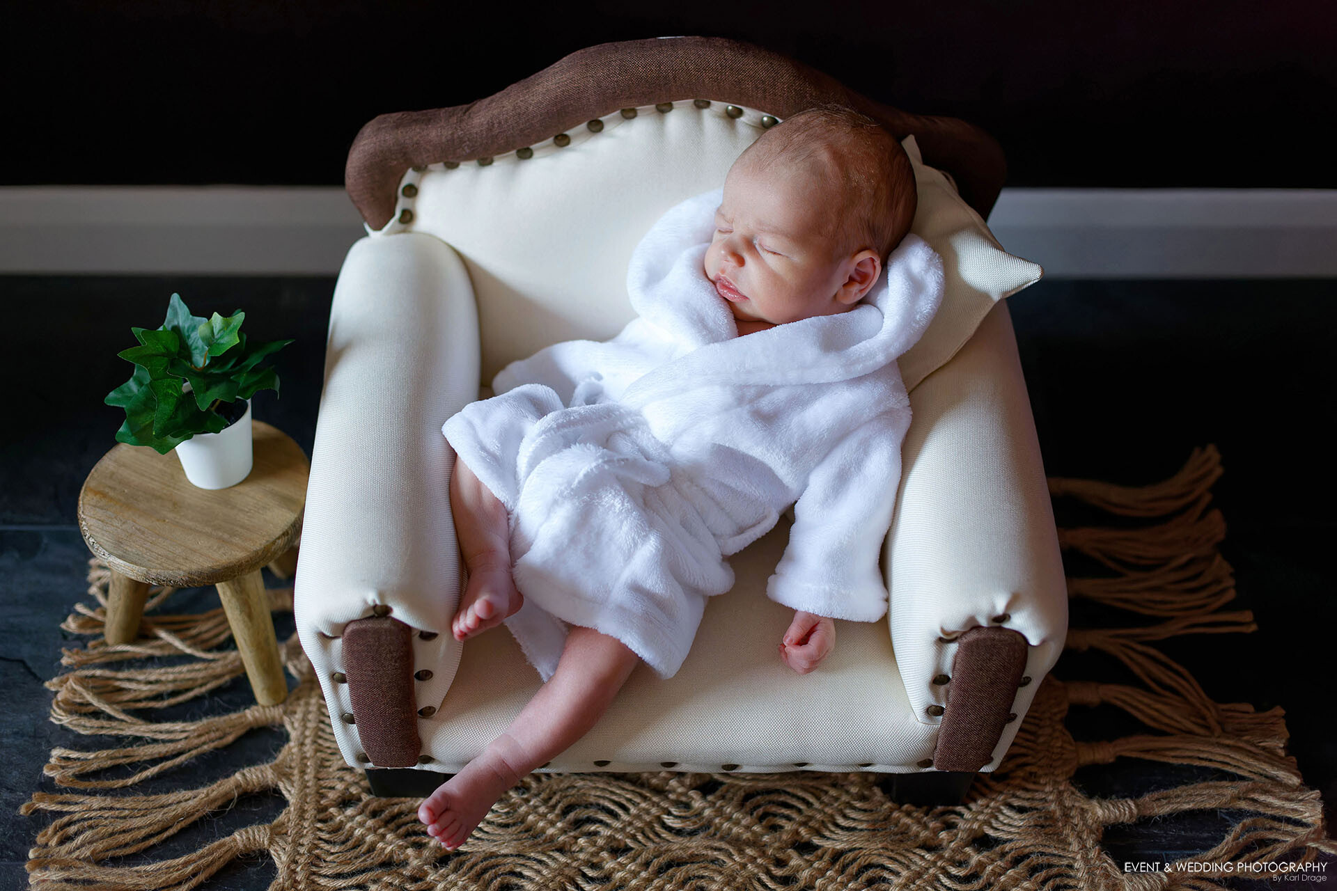 Newborn baby boy wearing a white dressing gown asleep on a white and brown upholstered sofa.