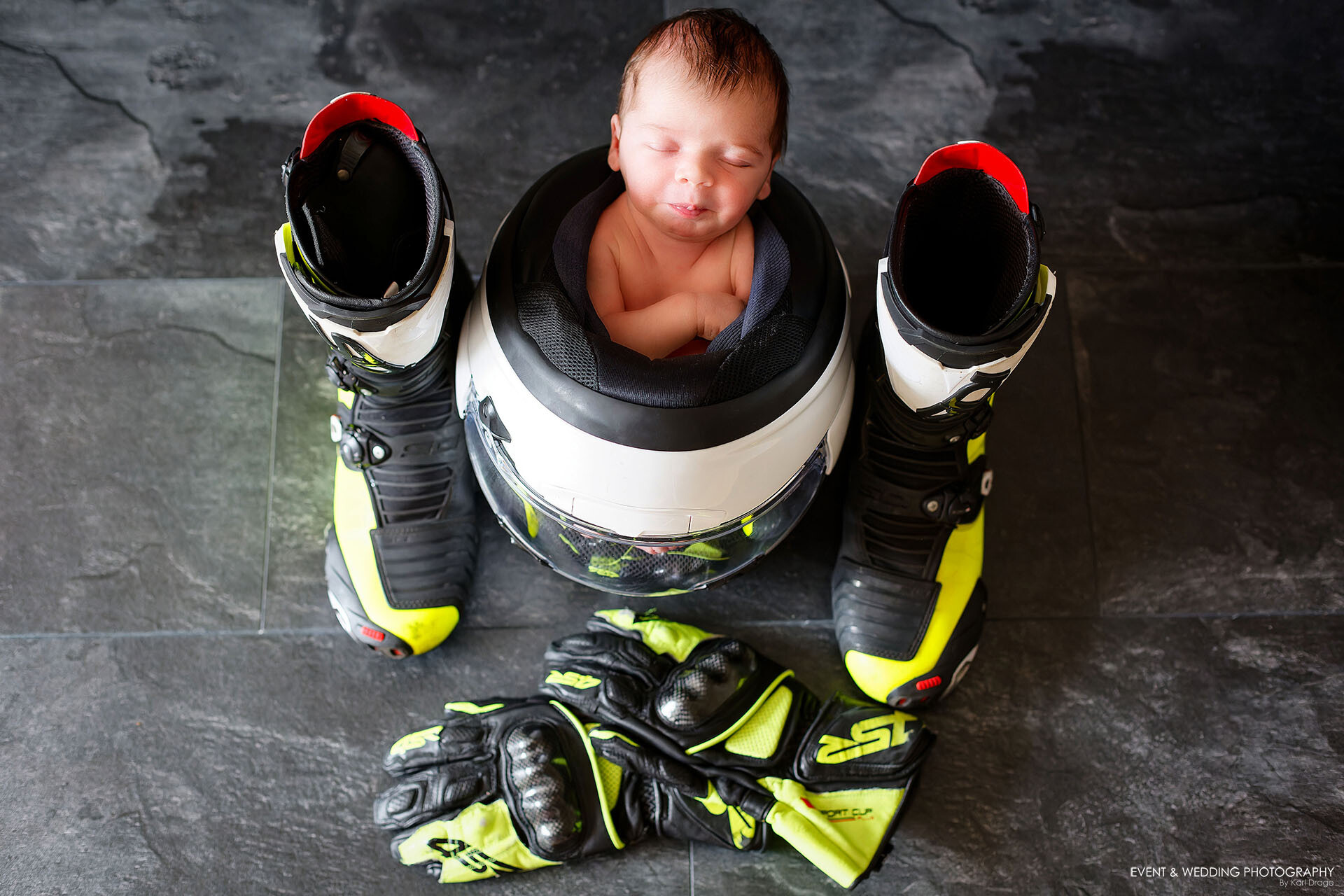 A newborn baby girl asleep in her daddy's upturned motorcycle racing helmet flanked by his boots and gloves
