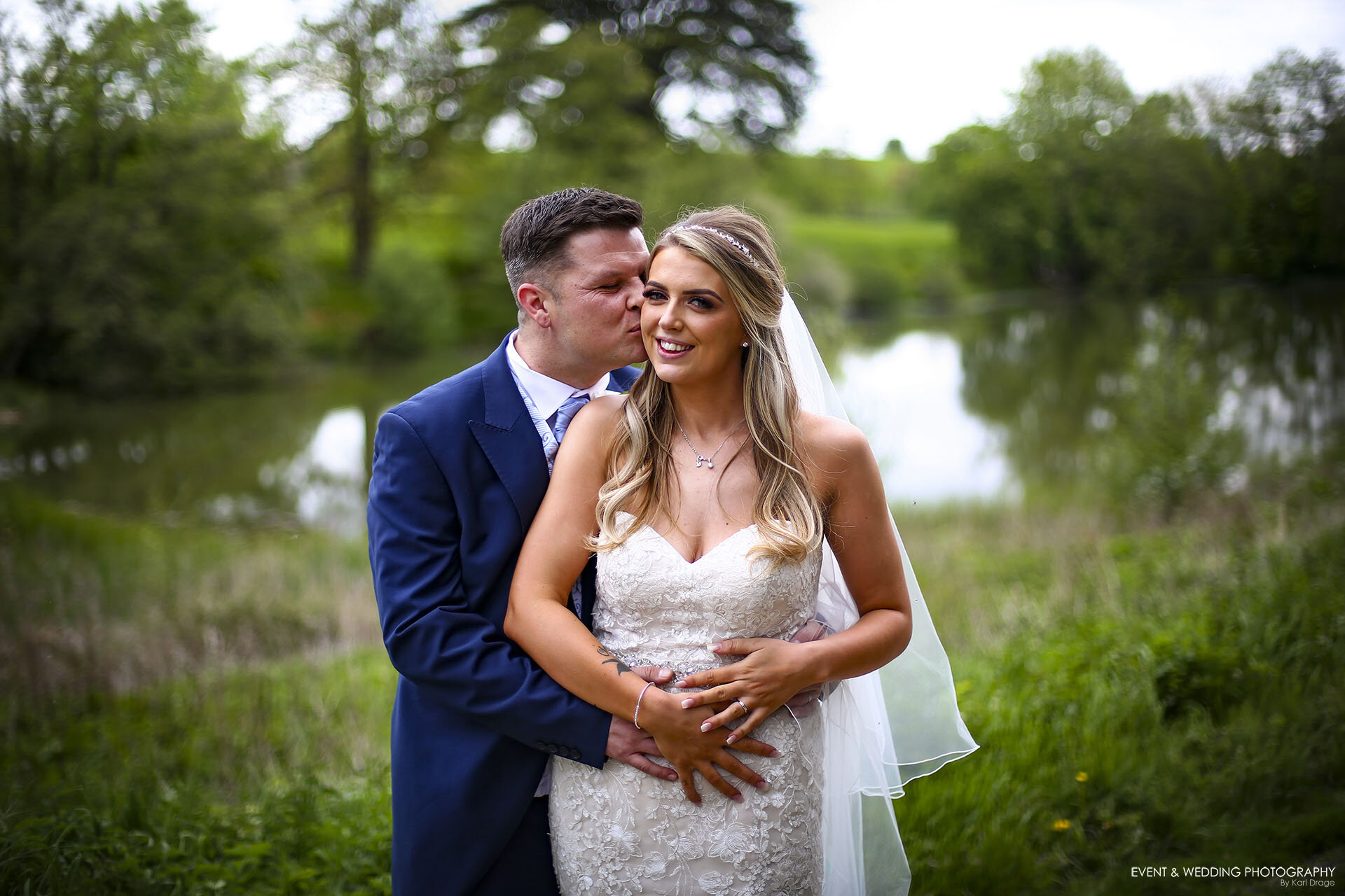A groom gives his new wife a peck on the cheek near the lake by Fawsley Hall on their wedding day