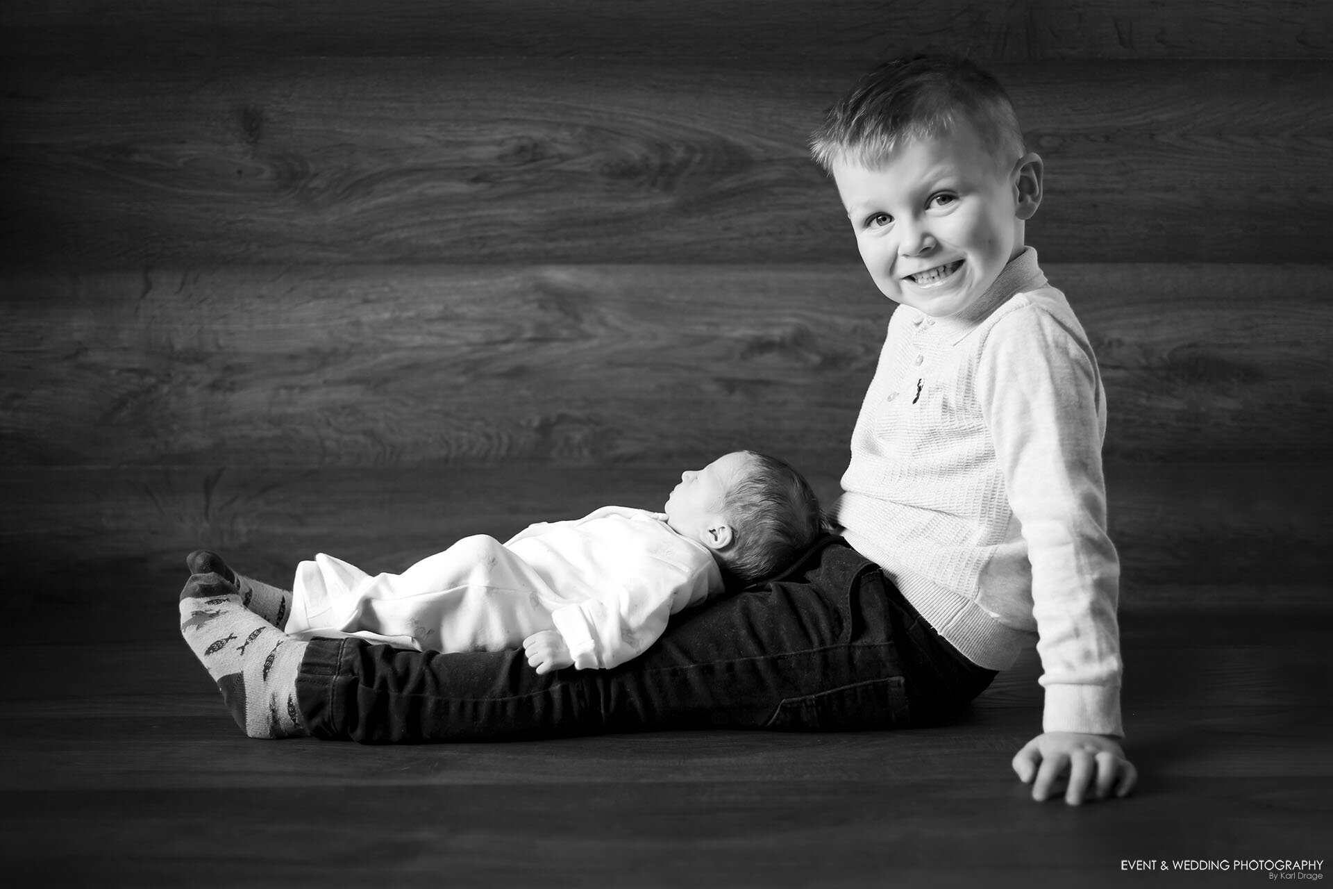 Black and white shot of a boy sitting with his newborn baby sister lying in between his outstretched legs