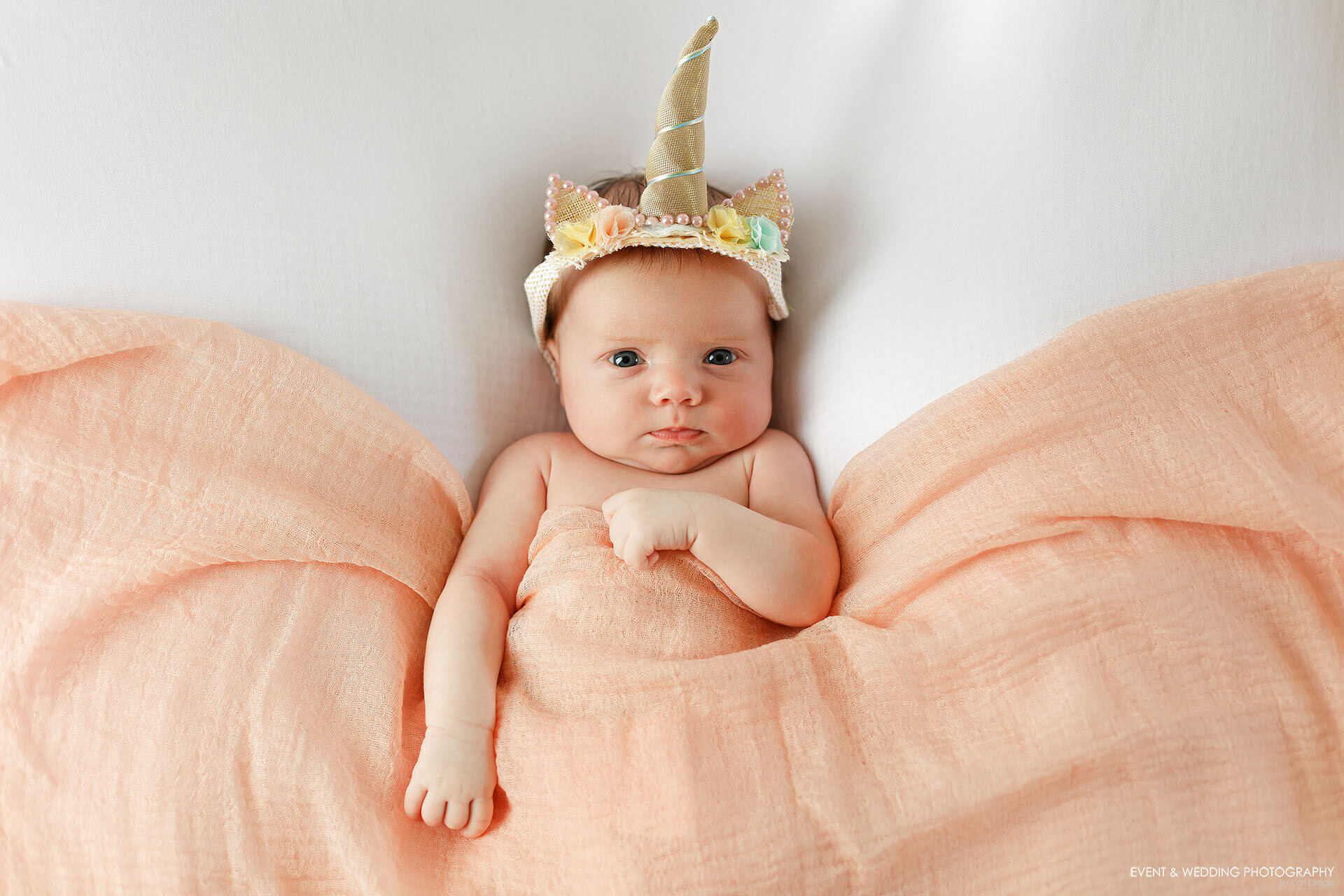 A newborn baby girl lays on a bean bag wearing a unicorn horn headband.