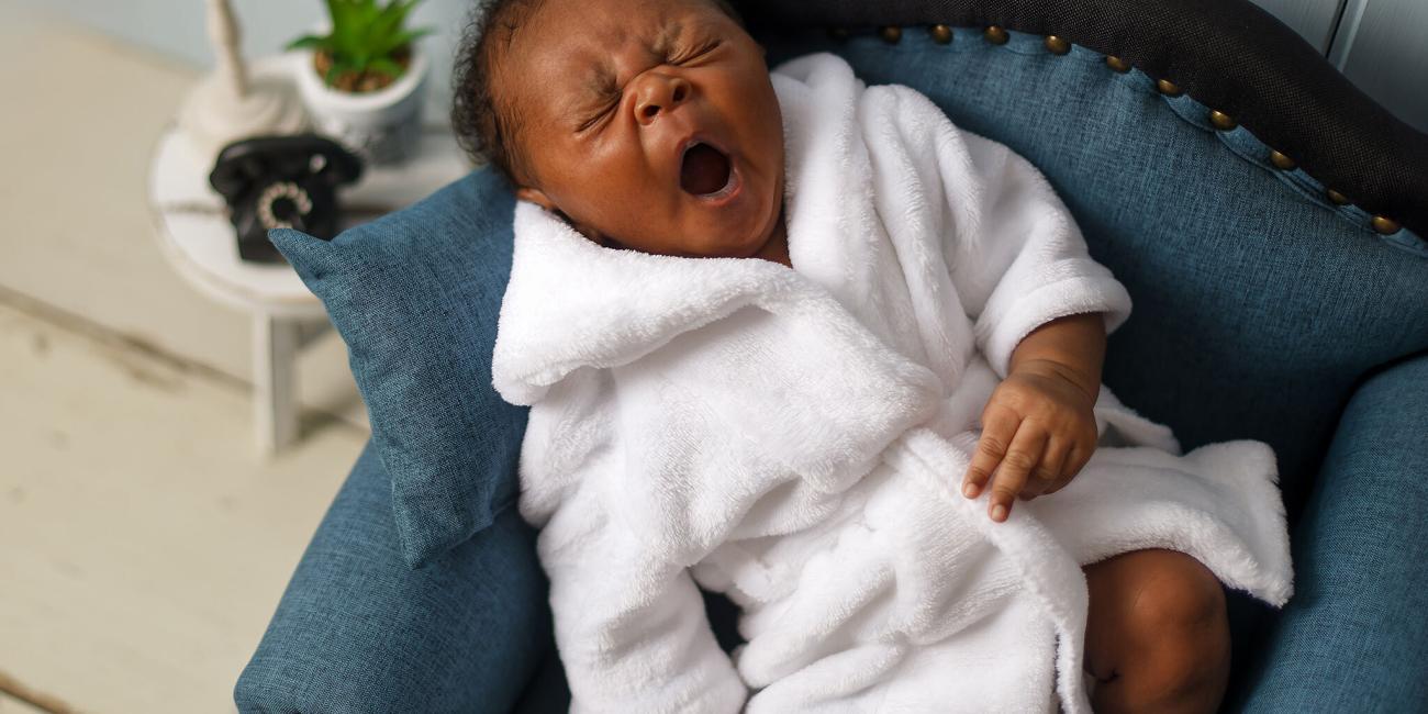 A newborn baby boy yawns in his blue arm chair, while wearing a white dressing gown at his Wellingborough newborn photo shoot