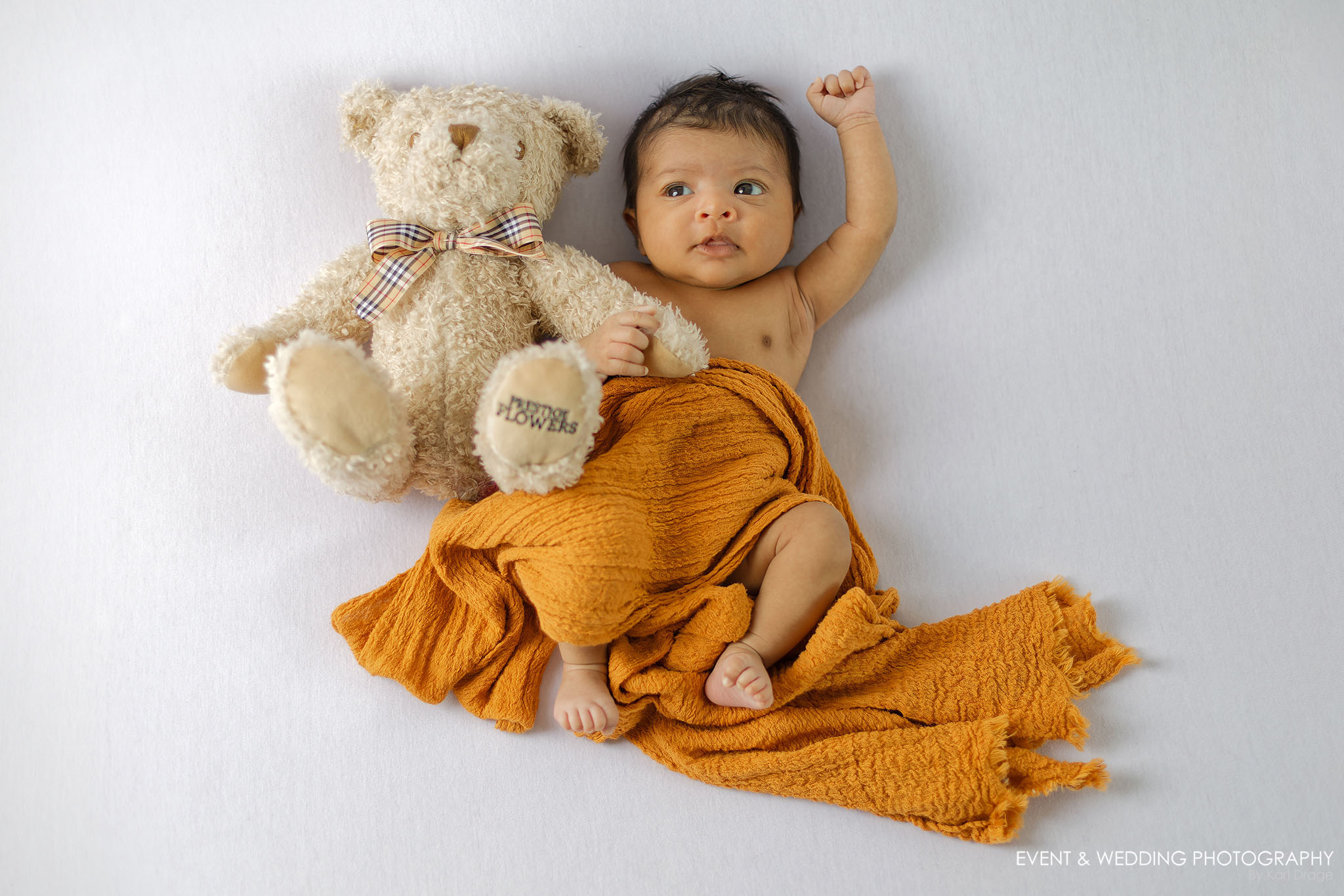 An open-eyed newborn baby girl waves her left arm as she hugs her teddy in her right.