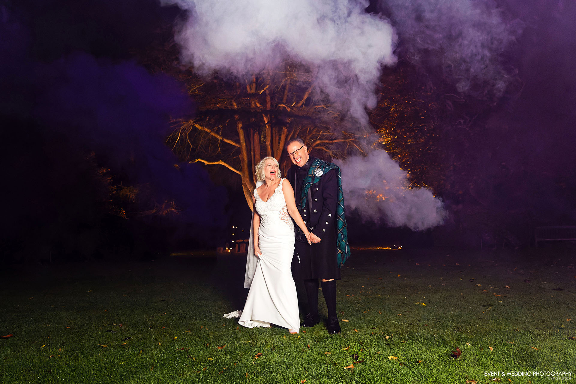 A bride and groom have their picture taken outside on the grass, in the dark, with purple and white smoke grenades billowing behind them.